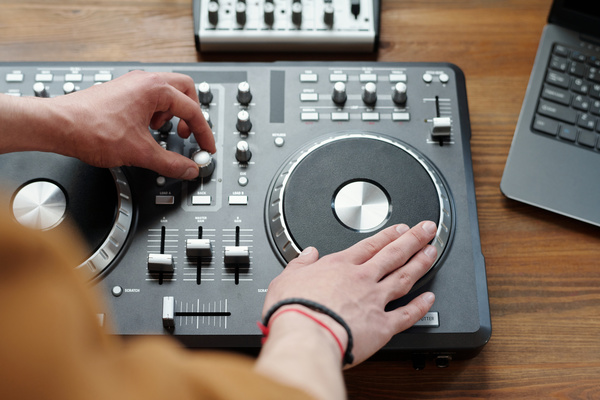 A guy with bracelets on his arm creates music by touching the disc and adjusting the equalizer on a black DJ console with silver elements which stands next to a laptop on a wooden table